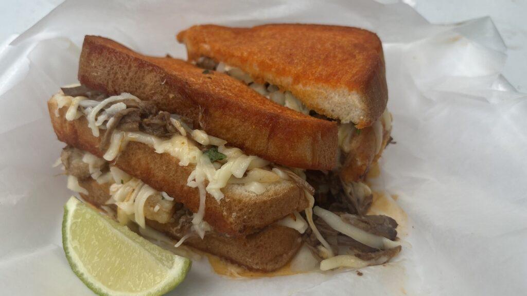 A grilled birria sandwich with melted cheese served with a lime wedge.
